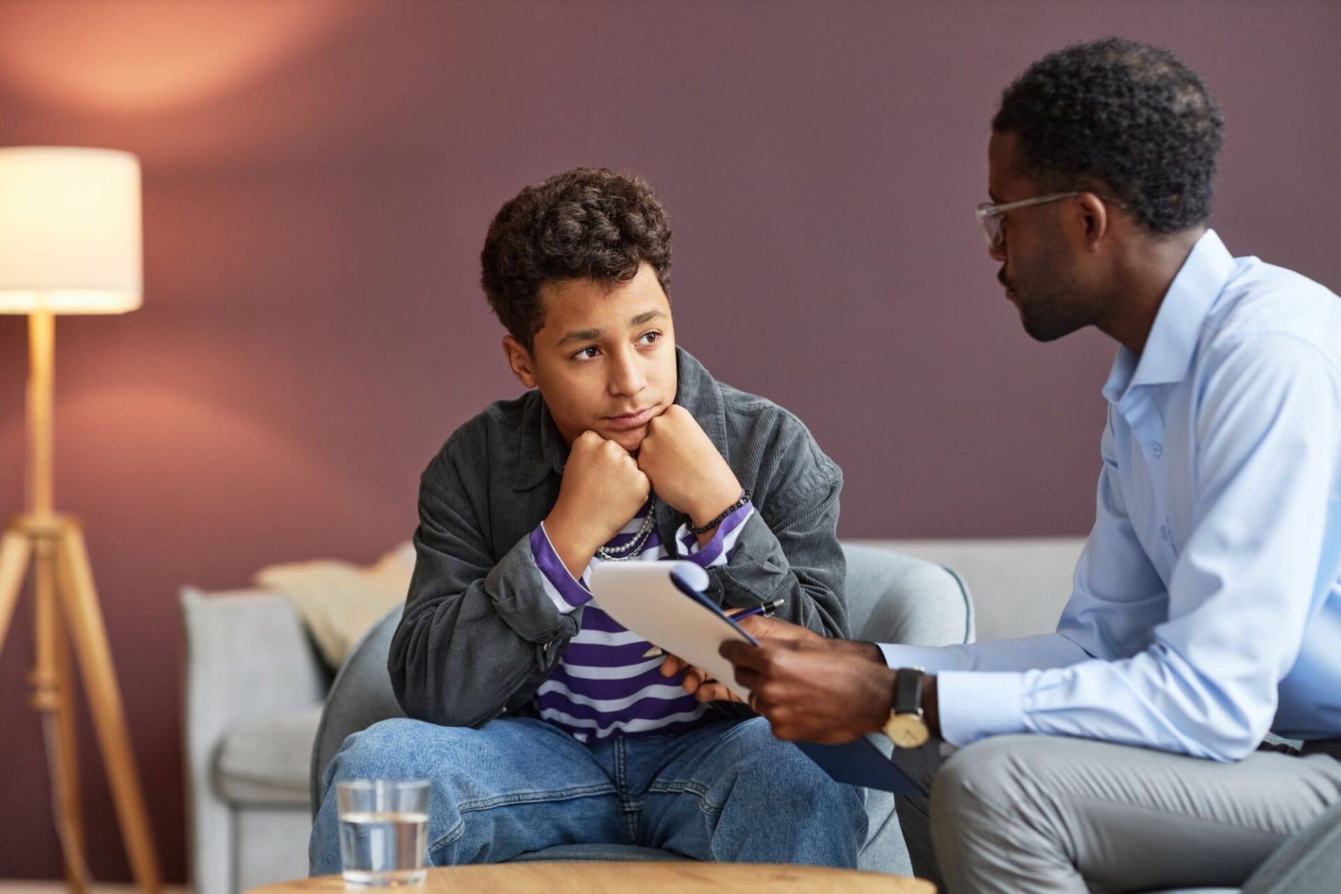 A teenage boy sits with his hands on his chin, listening to an adult man holding a notepad during a conversation in a living room setting.