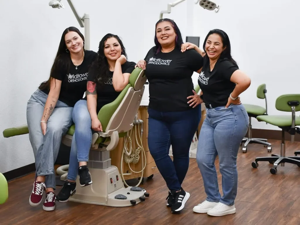 Four smiling staff members wearing black Wildflower Orthodontics t-shirts in a dental office.