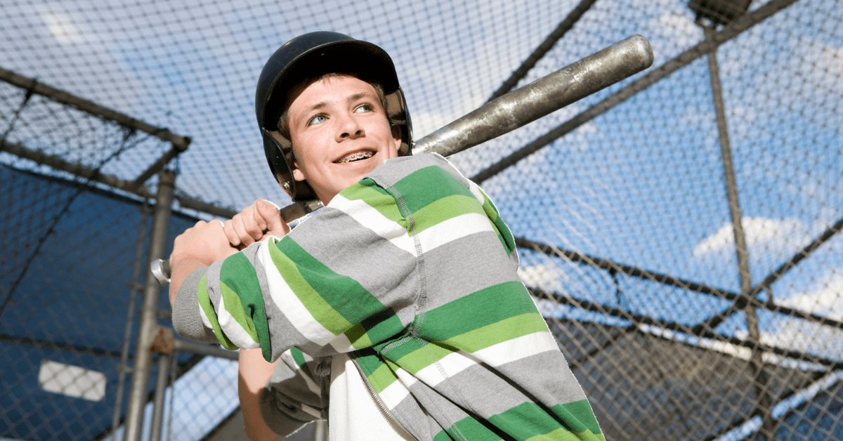 Teenager wearing a green striped hoodie and helmet holds a baseball bat, preparing to swing at a batting cage, confident with financing braces through easy braces payment plans.