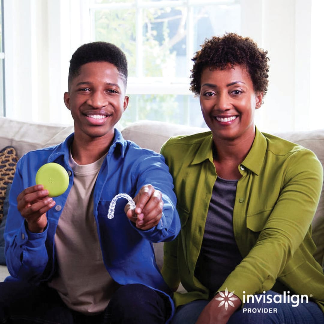 A smiling man holds a green Invisalign case and aligner tray while sitting next to a smiling woman on a couch in Round Rock; perfect smiles for teens and adults. Invisalign Provider logo in corner.