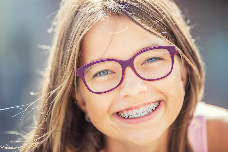Smiling child with long hair, wearing purple glasses and child braces from orthodontics, looking directly at the camera.