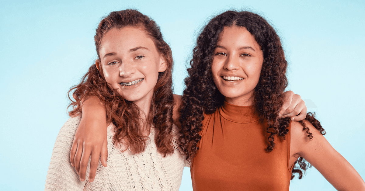 Two teenage girls stand side by side smiling, one with braces for kids and wearing a white sweater, the other in a sleeveless orange top with her arm around her friend’s shoulders against a blue background.