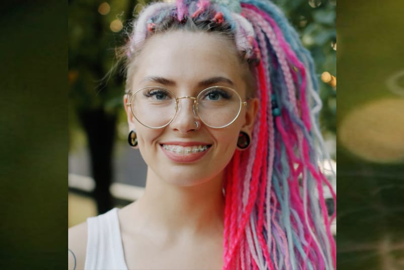A person with round glasses, Damon Clear braces, and colorful pink, blue, and white dreadlocks stands outdoors in Round Rock, smiling at the camera.