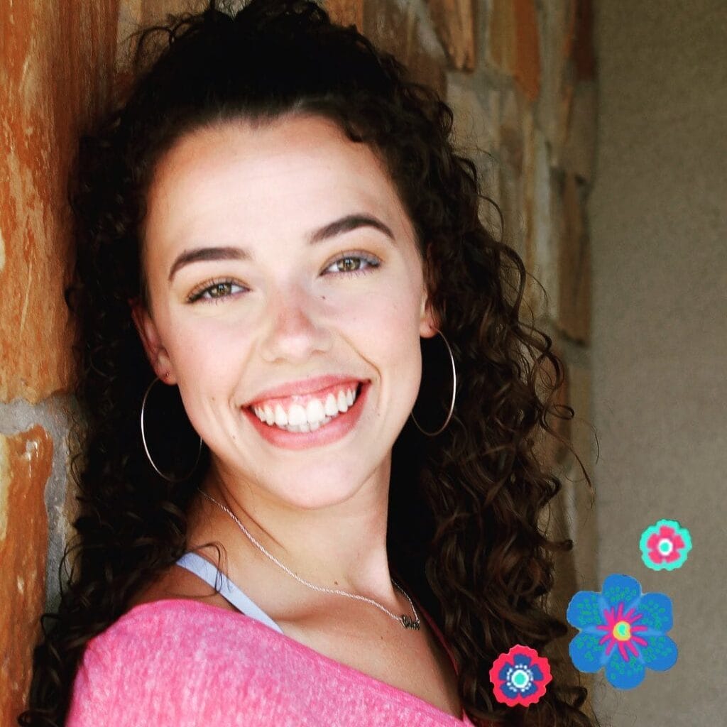 Young woman with curly hair and hoop earrings smiling in front of a stone wall, wearing a pink top with flower stickers added to the image corner.