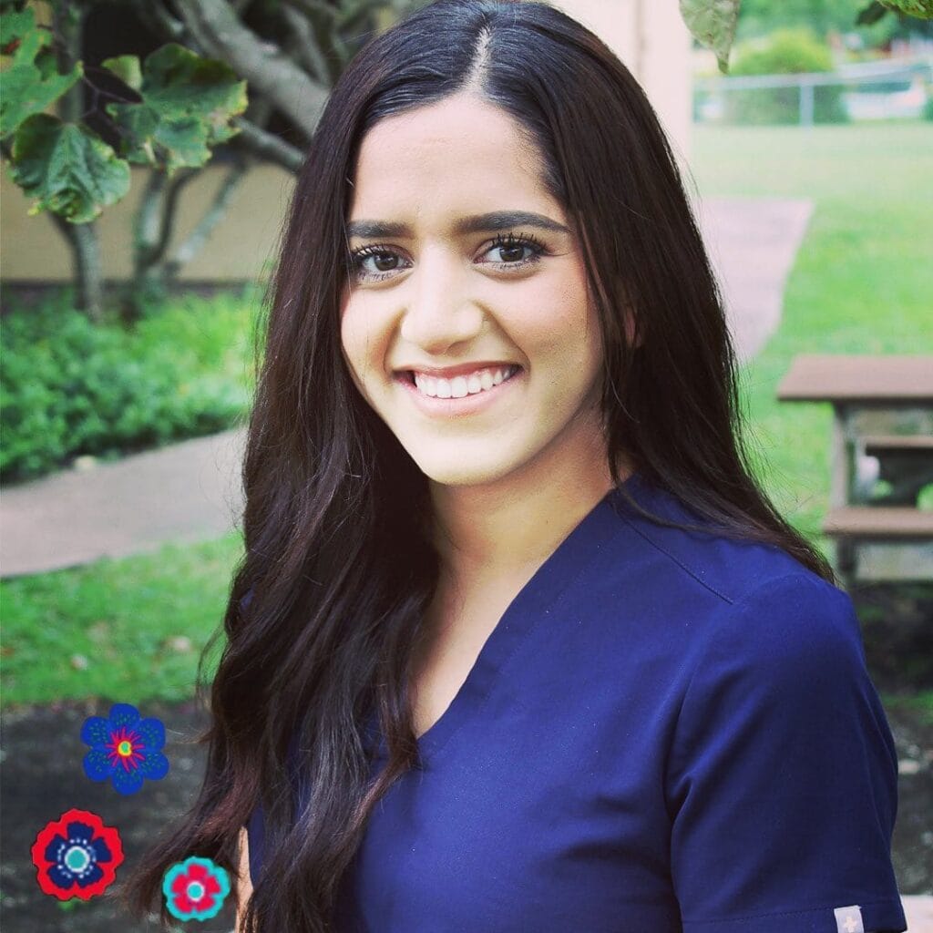 A woman with long dark hair wearing a navy blue top smiles outdoors near a picnic table and trees. Colorful flower graphics are in the bottom left corner.