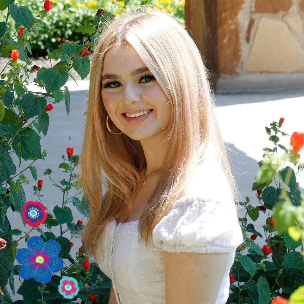 A young woman with long blonde hair wearing a white top stands outdoors near flowering plants, smiling at the camera.