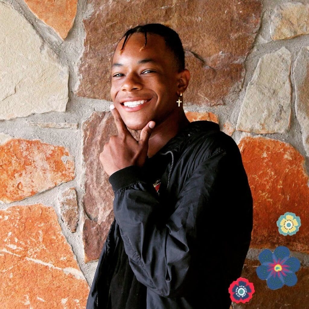 A smiling young person poses with a hand on their chin in front of a stone wall, wearing a black jacket and a cross earring. Three flower graphics are in the lower right corner.