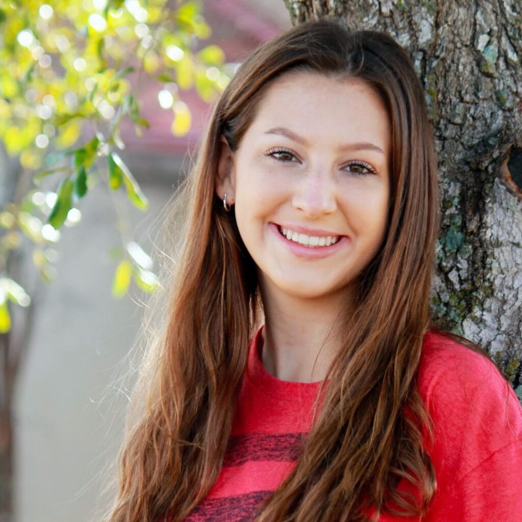 Young woman with long brown hair, wearing a red shirt, standing outdoors next to a tree and smiling at the camera.