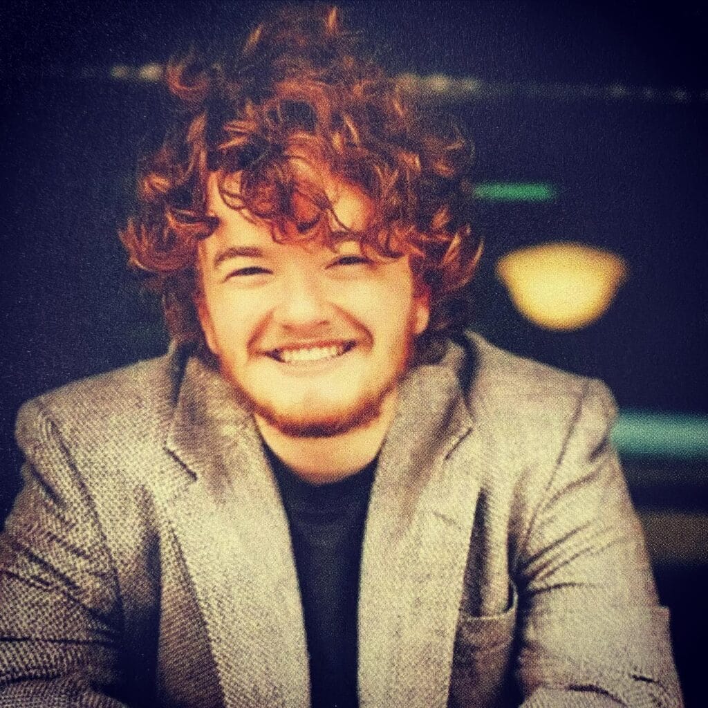 A person with curly red hair and a beard is smiling, wearing a gray blazer over a dark shirt, seated indoors with blurred lights in the background.