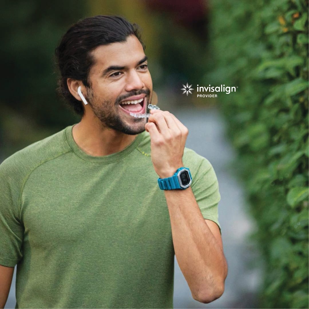 A man in a green shirt with wireless earbuds inserts an Invisalign aligner into his mouth while outdoors.
