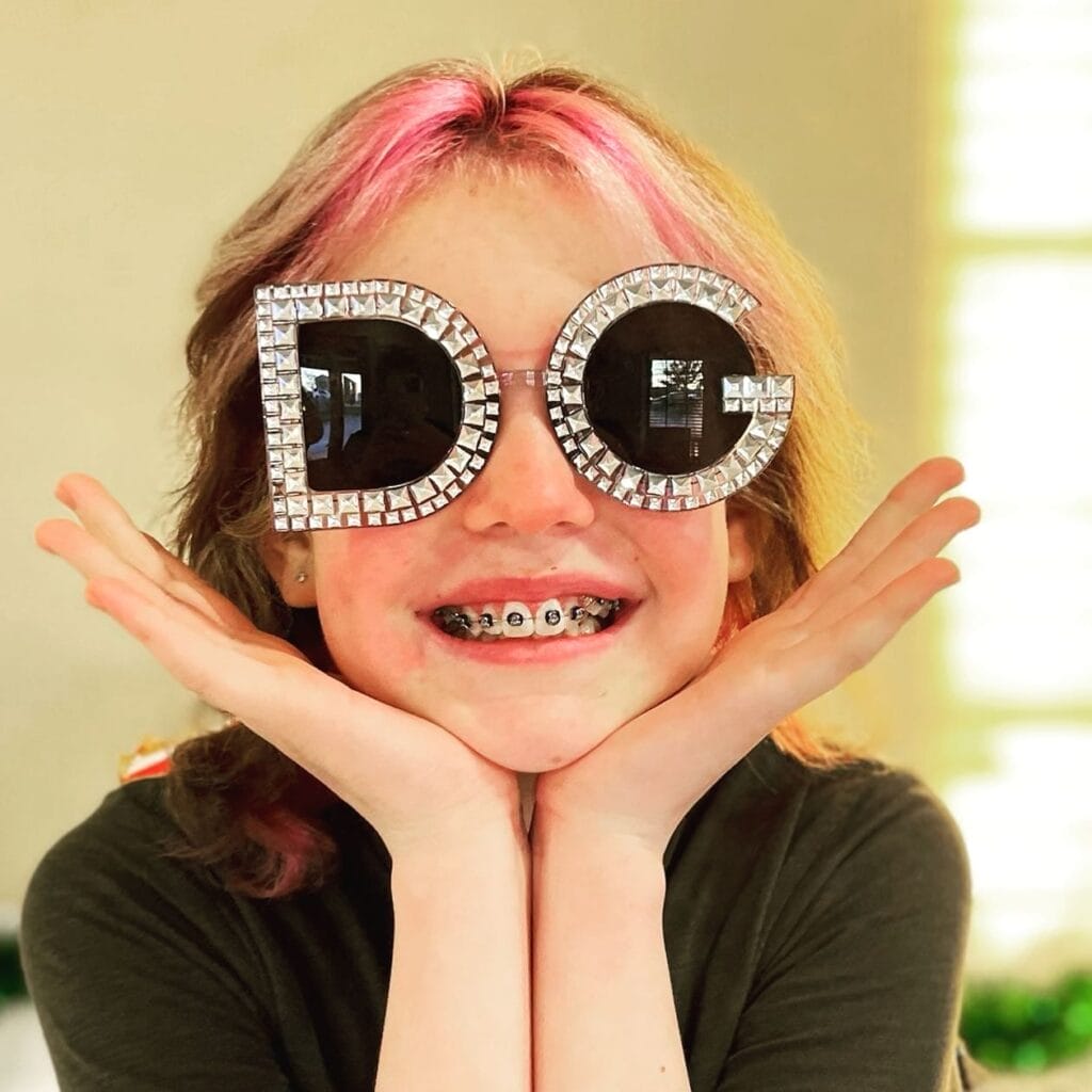 Smiling child with pink hair and braces wears oversized sunglasses with rhinestone letters "D" and "G," resting chin on hands.