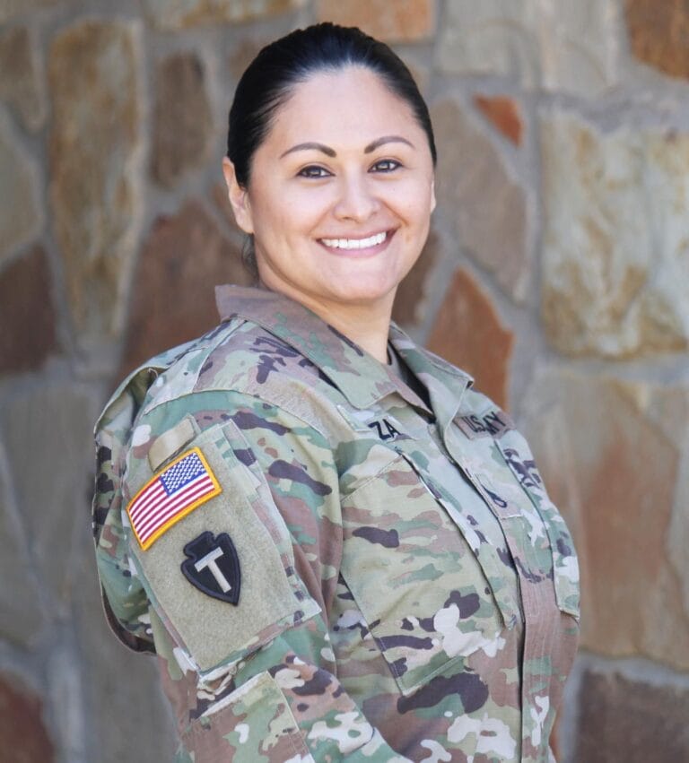 A person in a U.S. Army camouflage uniform smiles at the camera, standing in front of a stone wall. An American flag and a unit patch are visible on the uniform sleeve.