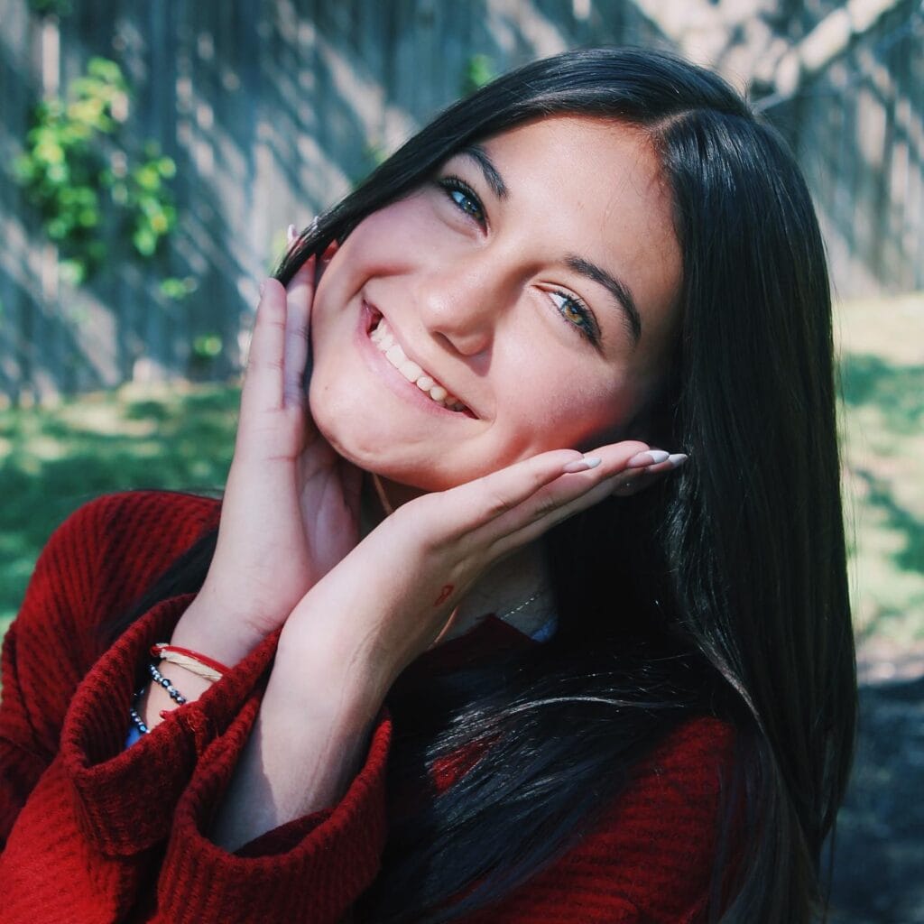 A young woman with long dark hair smiles outdoors, holding her face with both hands. She is wearing a red sweater and bracelets. A wooden fence and greenery are in the background.