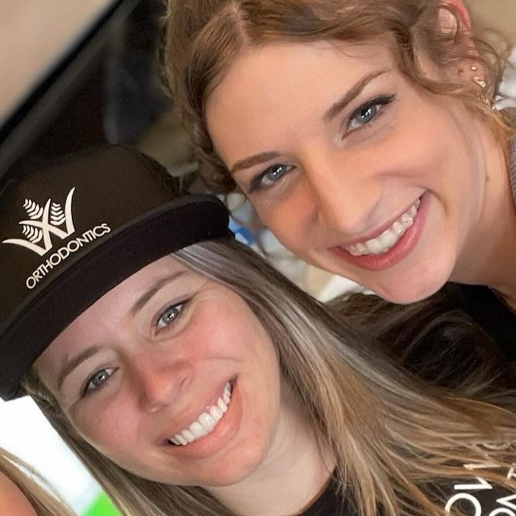 Two women smiling at the camera; one wears a black cap with a white logo and the word "ORTHODONTICS.