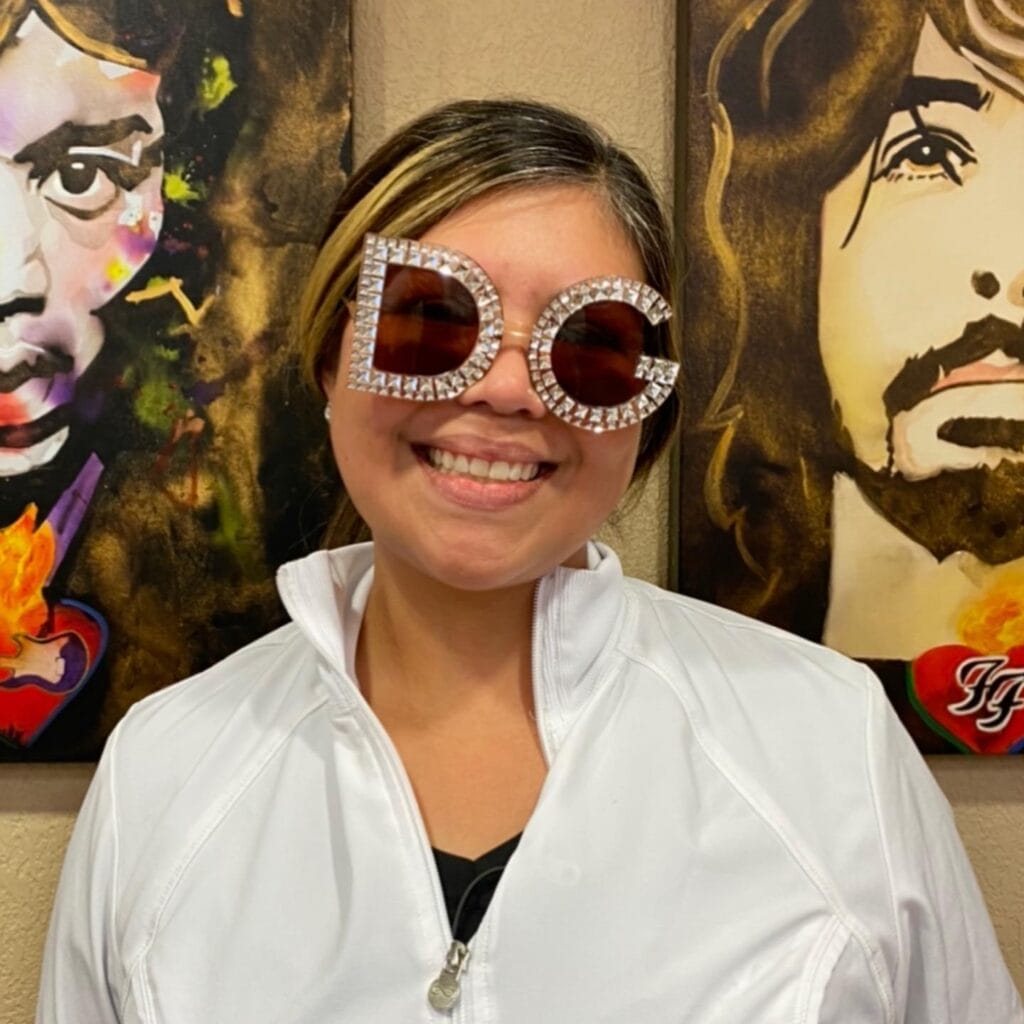 A woman in a white jacket smiles while wearing large, round sunglasses decorated with rhinestone "DG" letters. Two colorful, painted portraits are visible in the background.