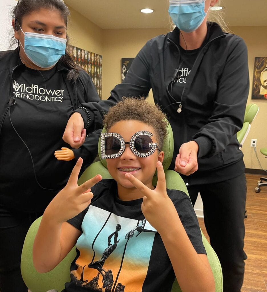 A child wearing large decorative glasses sits in a dental chair, flashing peace signs, with two masked orthodontic staff members standing beside him.