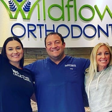 Three people stand smiling in front of a sign that reads "Wildflower Orthodontics." The person in the center wears blue scrubs, while the others wear casual tops.
