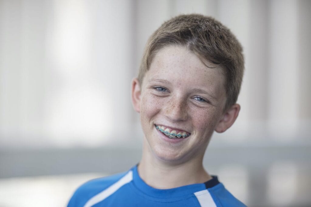 A boy with light brown hair and braces smiles while wearing a blue sports shirt, standing indoors with a blurred background.