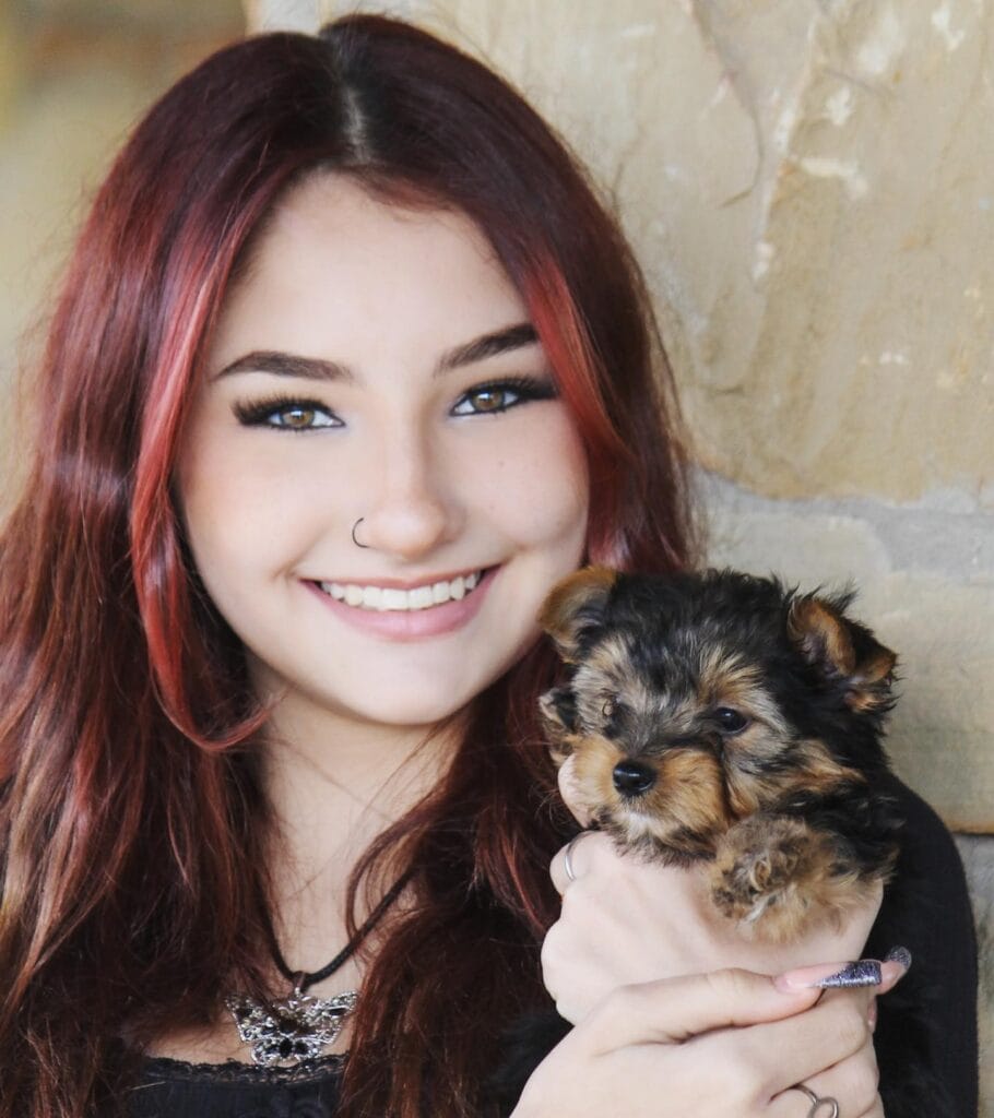 A young woman with long dark hair and a nose ring smiles while holding a small Yorkshire Terrier puppy in front of a stone wall.