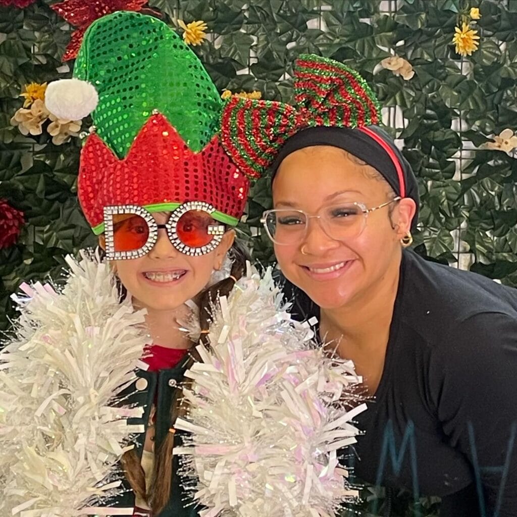 A child wearing a festive elf hat, large "DC" glasses, and holding white tinsel stands next to an adult in striped Christmas cat ears, both smiling at the camera.
