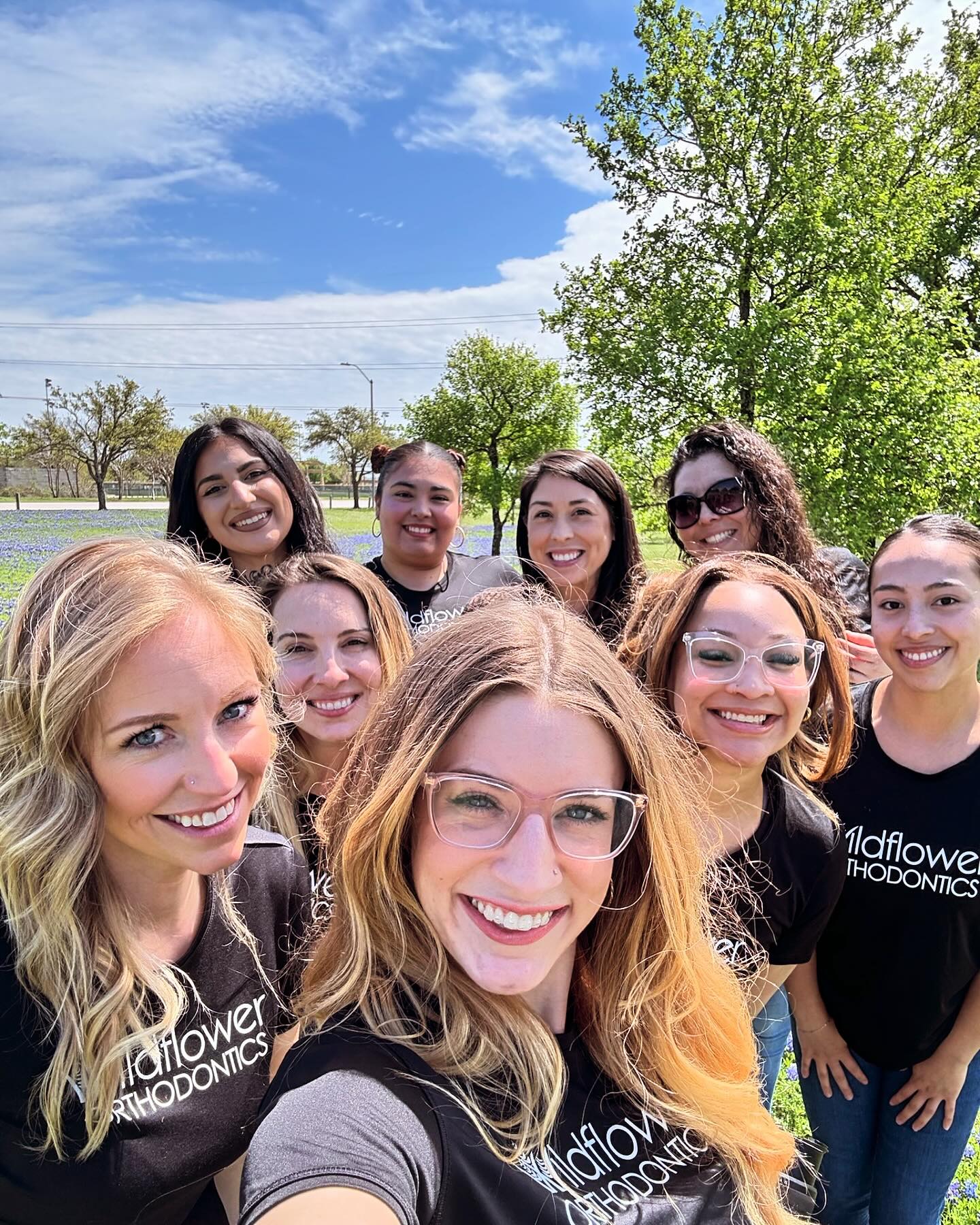 A group of nine women wearing "Wildflower Orthodontics" shirts pose outdoors in front of green trees and a partly cloudy sky.