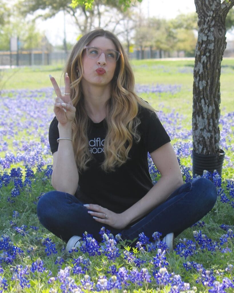 A woman with long, wavy hair sits cross-legged on grass among blue flowers, making a peace sign with her hand and puckering her lips. Trees and a fence are visible in the background.