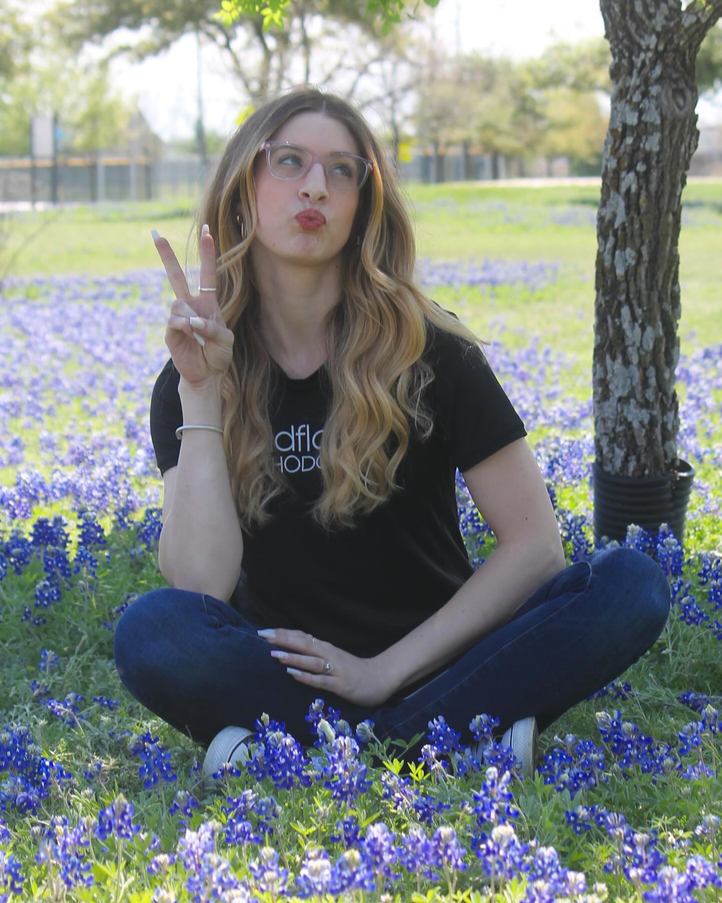 A woman with long, wavy hair sits cross-legged on grass among blue flowers, making a peace sign with her hand and puckering her lips. Trees and a fence are visible in the background.