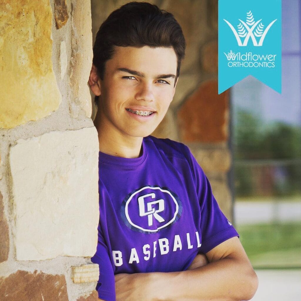 Teen boy with braces wearing a purple baseball shirt stands by a stone wall, smiling. Wildflower Orthodontics logo is visible in the upper right corner.