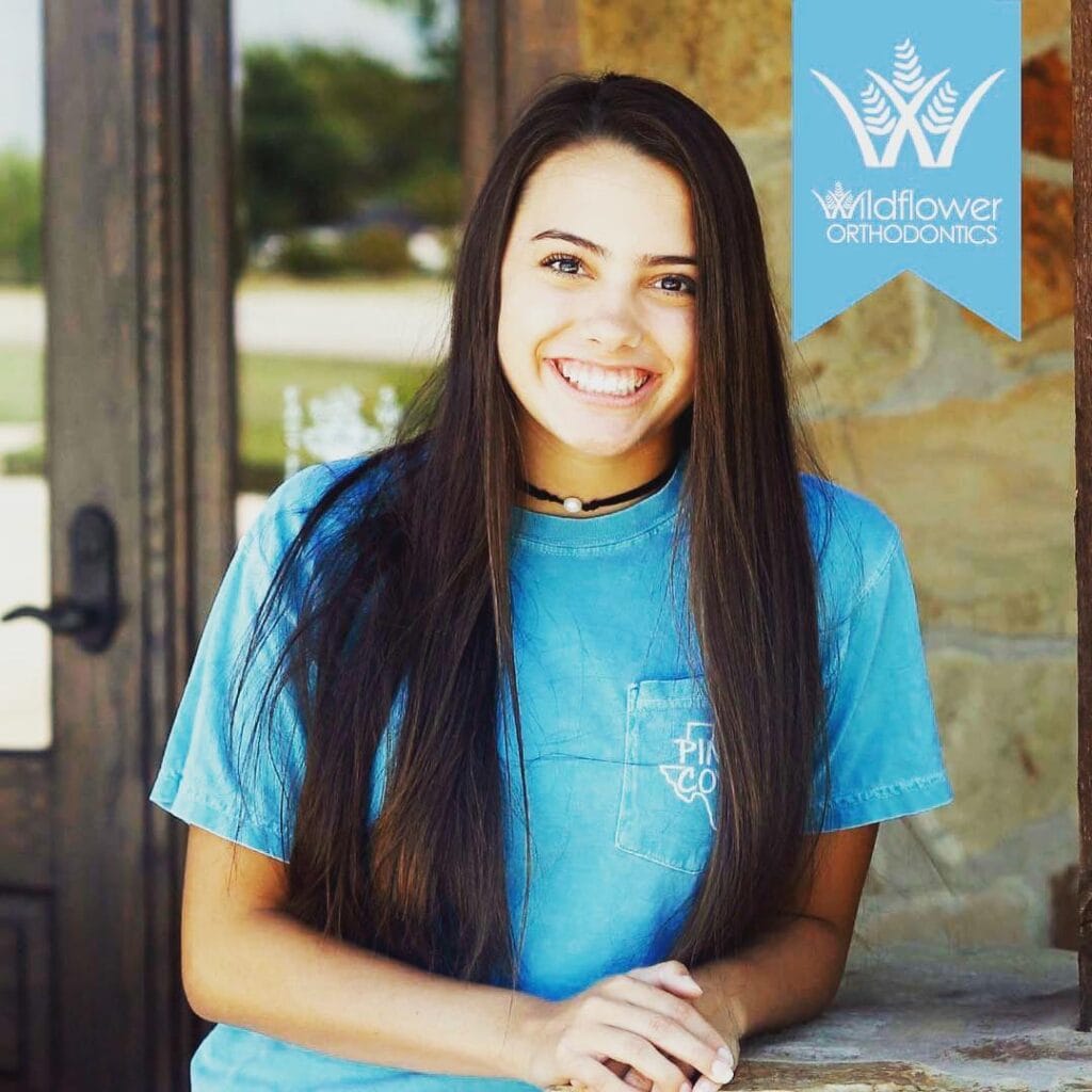 A young woman with long dark hair smiles at the camera, standing outside near a door. The Wildflower Orthodontics logo appears in the top right corner.