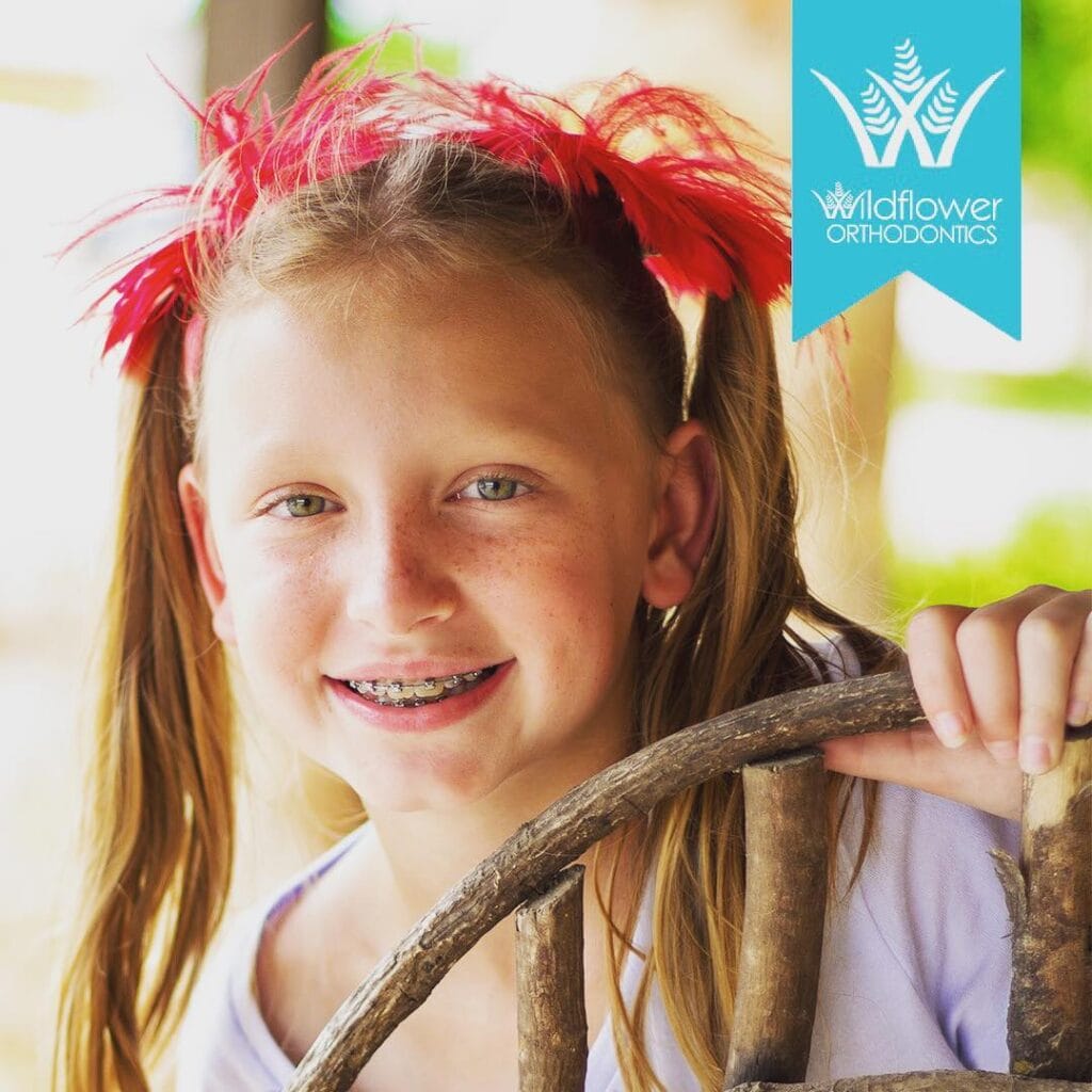 Smiling girl with braces and red feather hair accessories holds a wooden wagon wheel. Wildflower Orthodontics logo in top right corner.