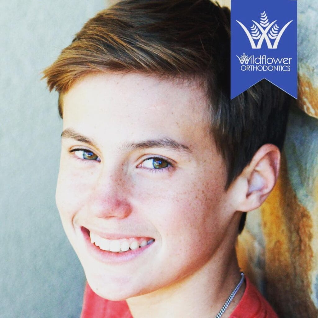 Teenager smiling at the camera, standing near a stone wall with a "Wildflower Orthodontics" logo in the top right corner.