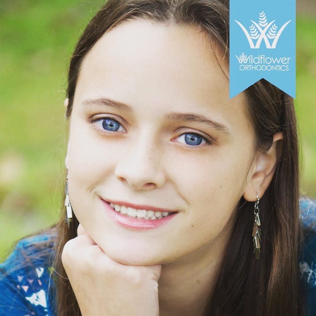 A young person with long brown hair and blue eyes smiles at the camera, resting their chin on their hand. A "Wildflower Orthodontics" logo appears in the top right corner.