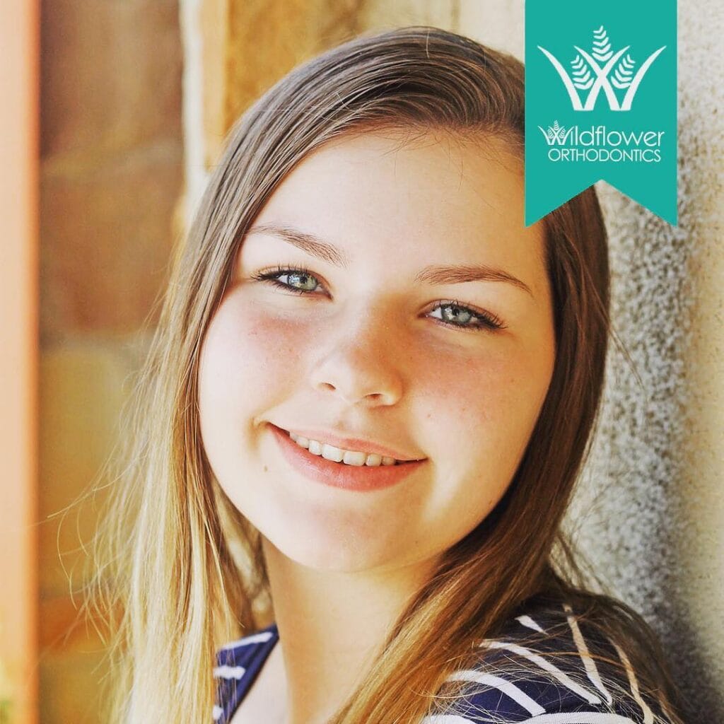 Teenage girl with long, straight hair smiling in front of a textured wall; Wildflower Orthodontics logo in the top right corner.