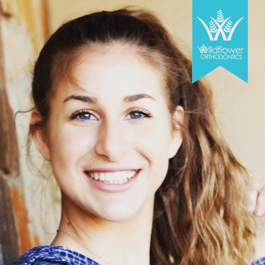 A young woman with long brown hair in a ponytail smiles at the camera; a "Wildflower Orthodontics" logo appears in the top right corner.