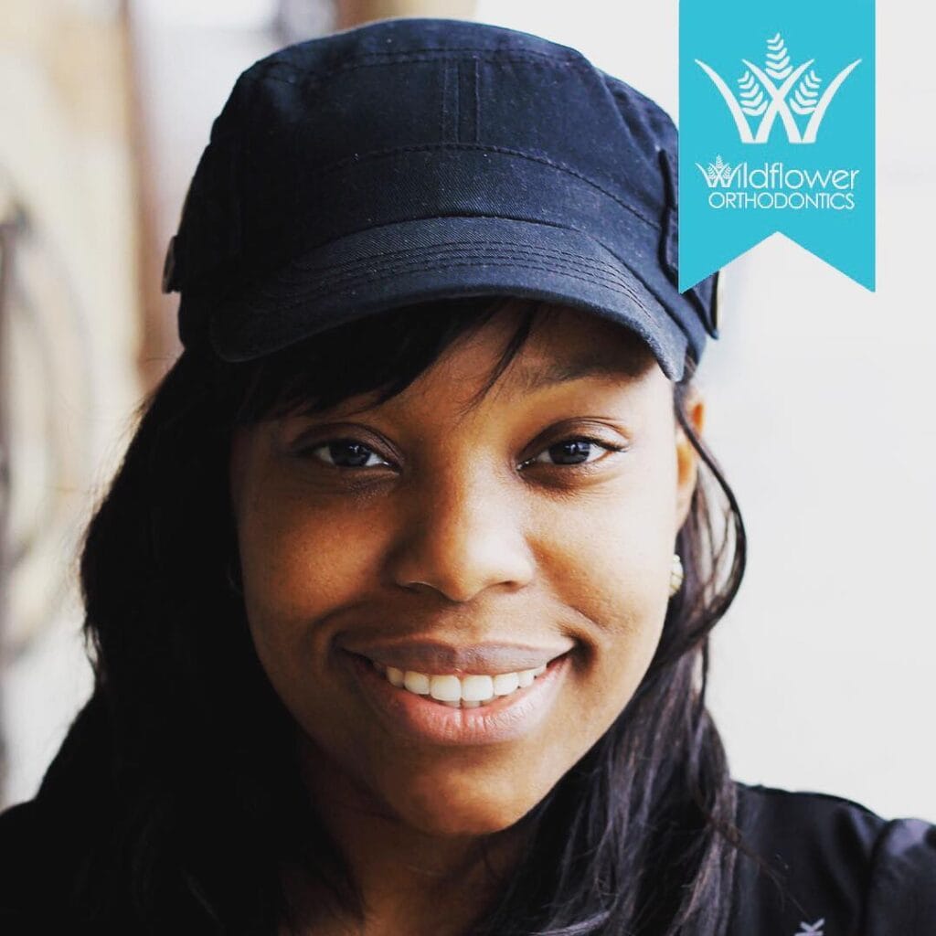 A woman wearing a dark cap smiles at the camera. In the upper right corner, there is a blue Wildflower Orthodontics logo.