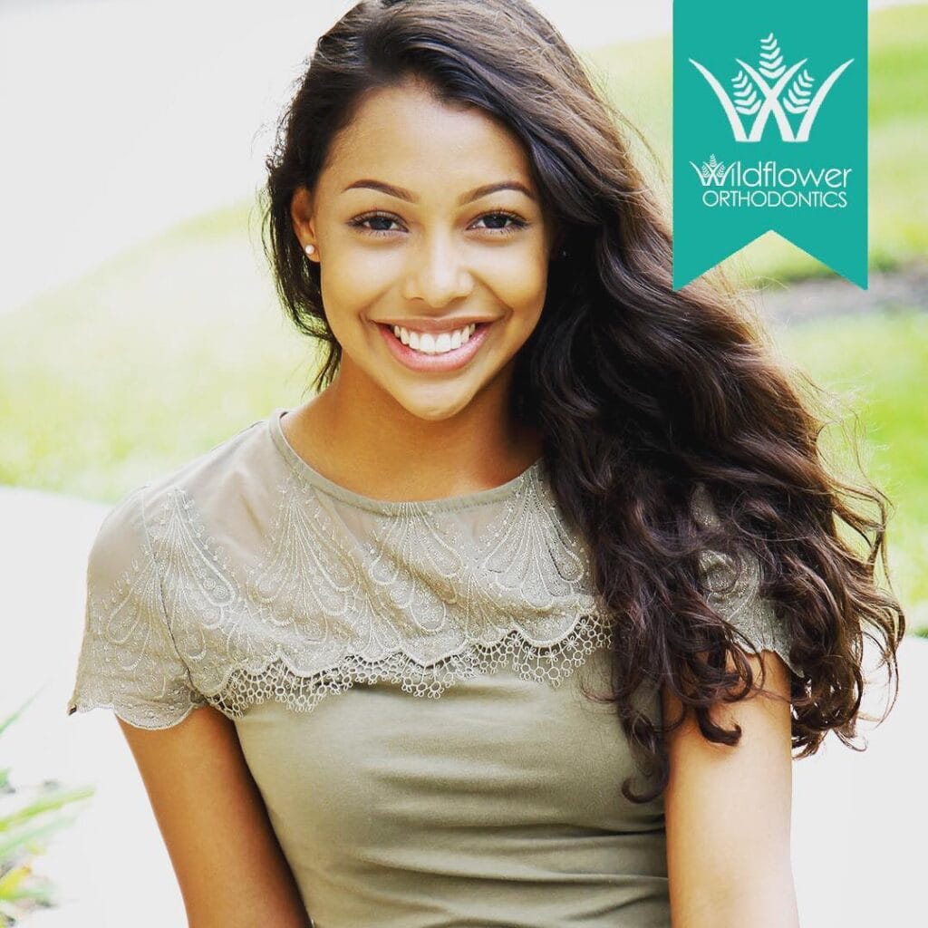 A young woman with long dark hair smiles at the camera. She is wearing a light green lace top. "Wildflower Orthodontics" logo appears in the top right corner.