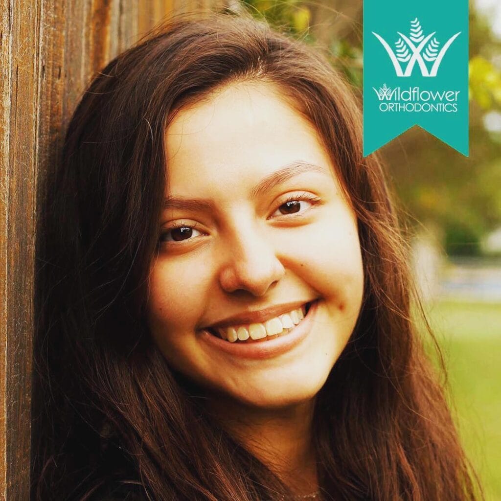 A young woman with long brown hair smiles near a wooden fence. The Wildflower Orthodontics logo appears in the top right corner.