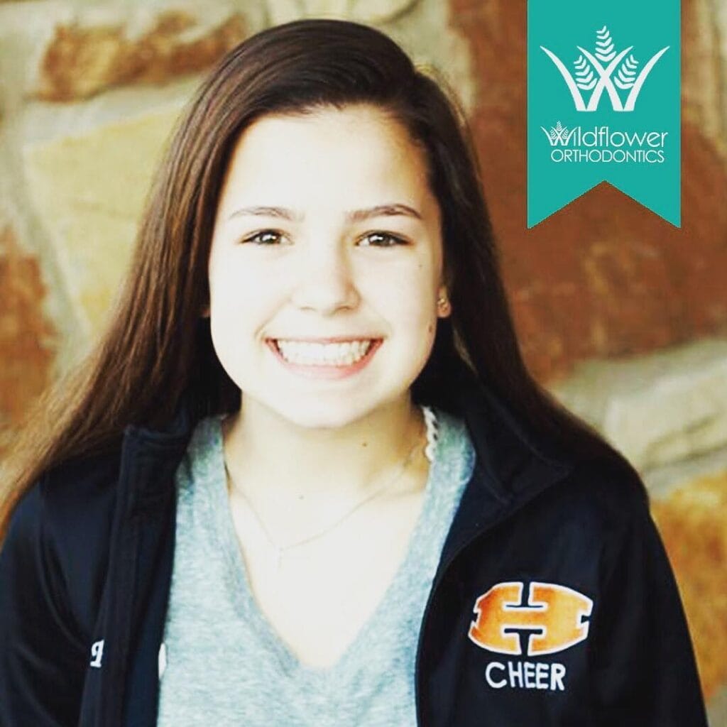 Smiling girl with long brown hair wearing a black "H Cheer" jacket, standing in front of a stone wall. Wildflower Orthodontics logo is in the upper right corner.