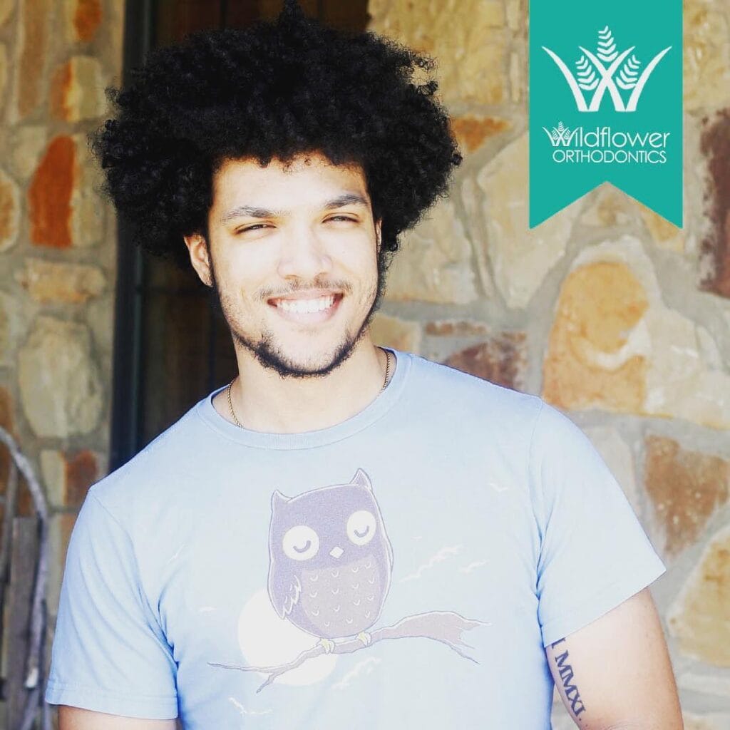 A smiling person with curly hair wears a light blue t-shirt with an owl graphic, standing in front of a stone wall. Wildflower Orthodontics logo appears in the top right corner.