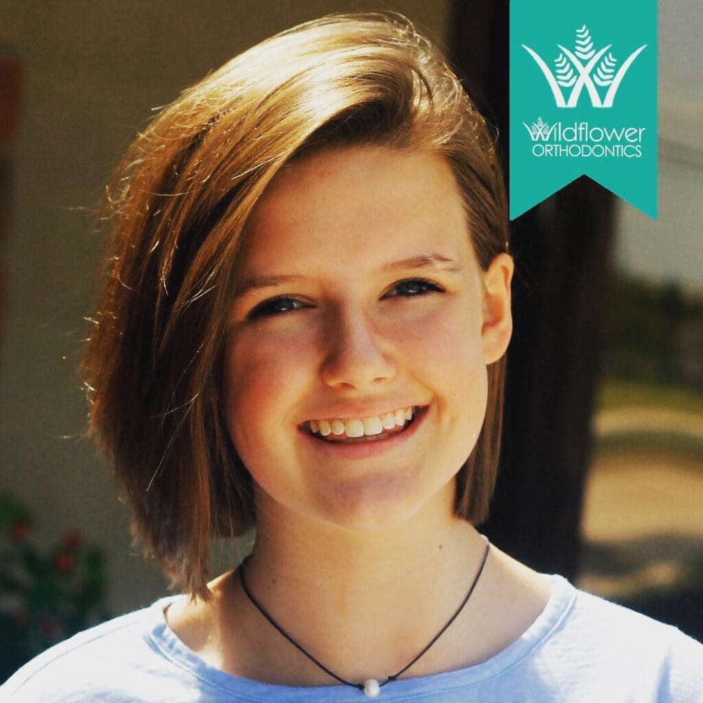 A smiling young person with short, light brown hair stands outdoors, wearing a light blue top and necklace. A "Wildflower Orthodontics" logo appears in the upper right corner.