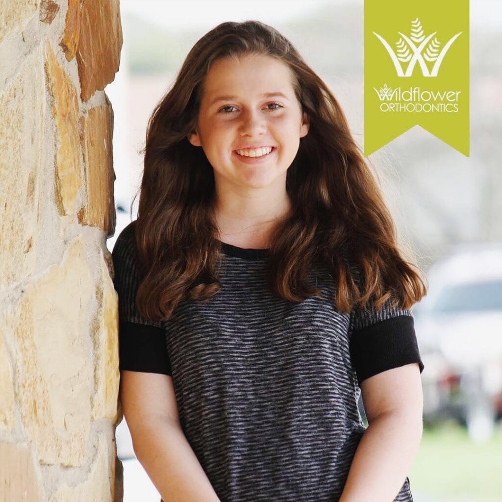 A young woman with long brown hair stands smiling near a stone wall. The Wildflower Orthodontics logo is visible in the upper right corner.