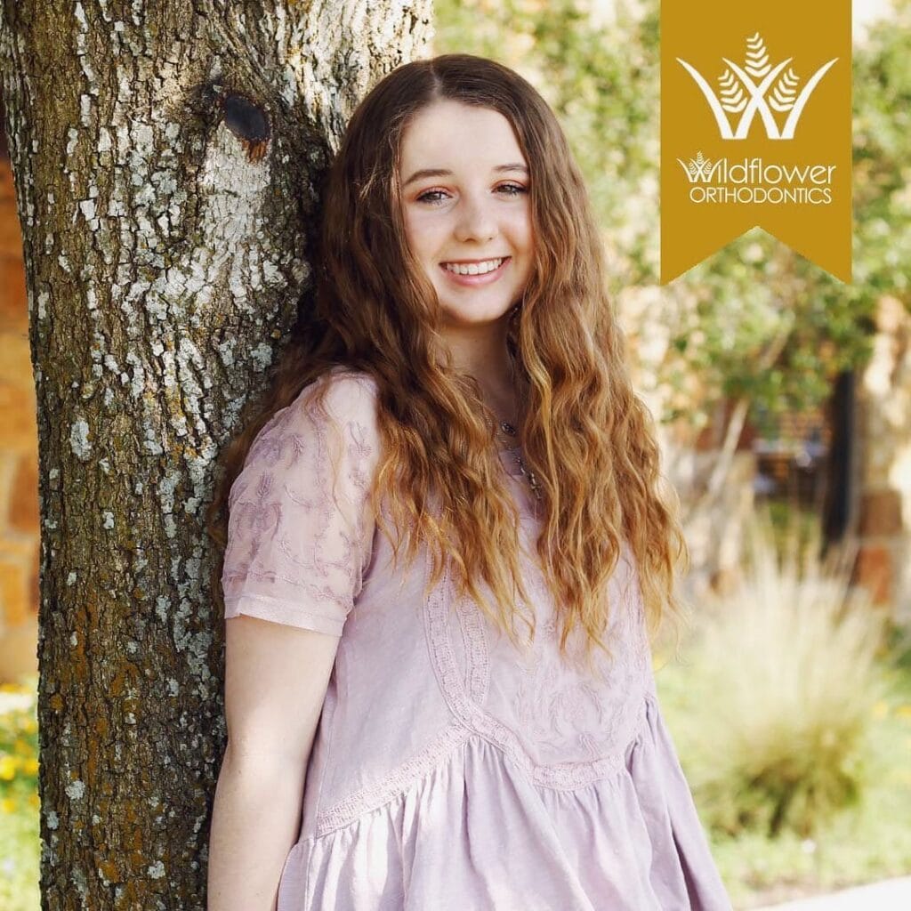 A young woman with long wavy hair in a light pink dress stands by a tree. The Wildflower Orthodontics logo is in the top right corner.