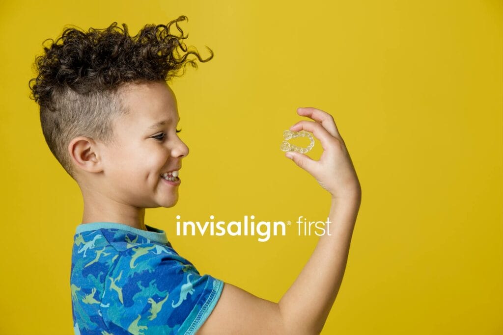 A young boy in a blue shirt smiles while holding a clear aligner with the words "invisalign first" overlaid on a yellow background.