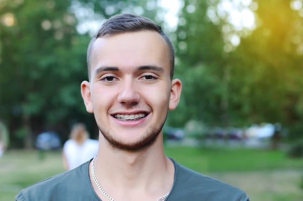 A young man with short hair and braces smiles at the camera outdoors, with a blurred green background.