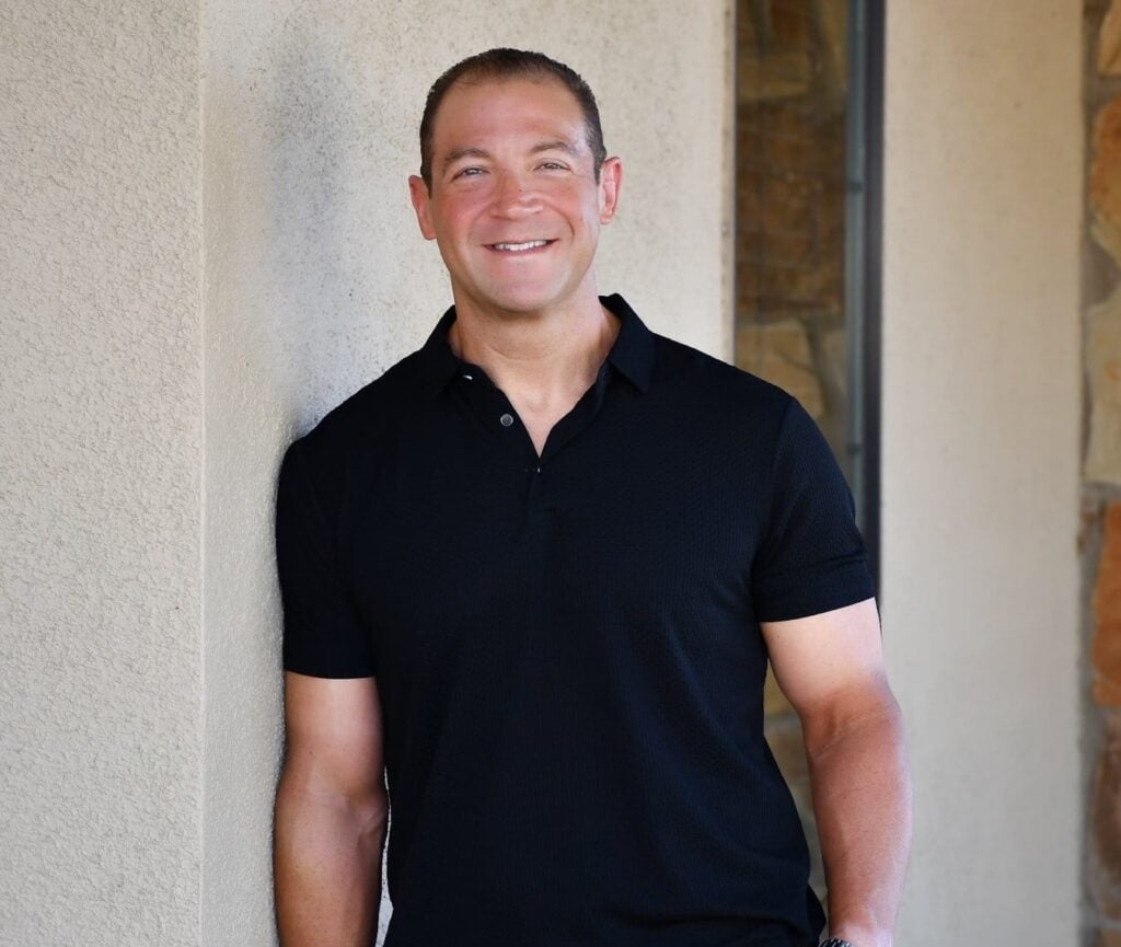 Dr. Joseph Giasi stands and smiles at the camera, wearing a black short-sleeve polo shirt, gray pants, and a watch, leaning against a beige wall.