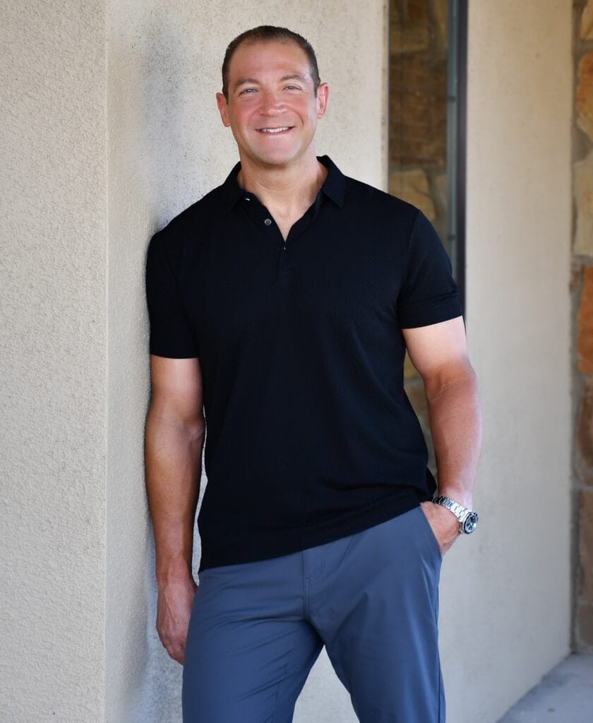 A smiling orthodontist stands against a light-colored wall, wearing a black polo shirt, gray pants, and a watch, with one hand in his pocket.