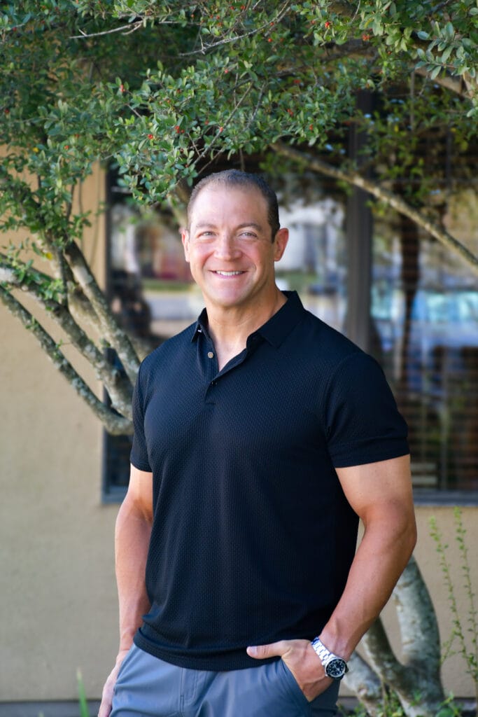 Dr. Joseph Giasi, wearing a black polo shirt and gray pants, stands outdoors in front of a building and wildflower greenery, smiling at the camera for a meet the doctor profile.