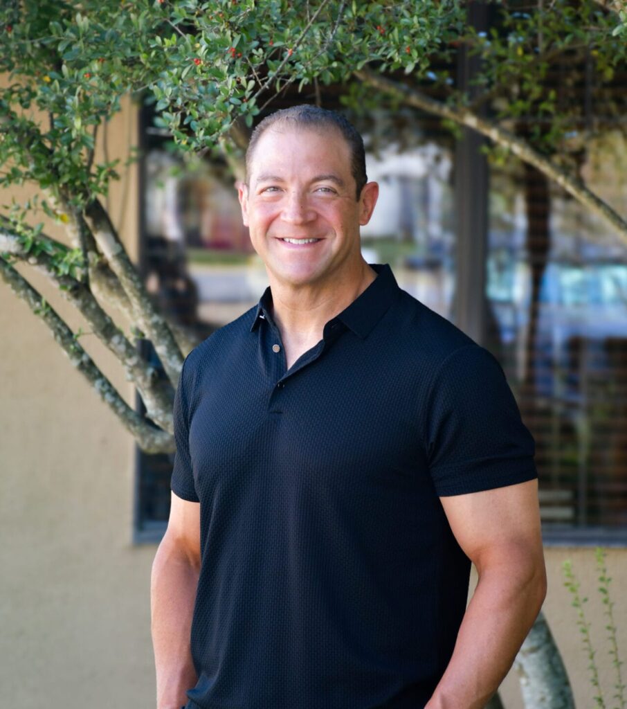 Dr. Joseph Giasi, wearing a black polo shirt and gray pants, stands outdoors in front of a building and wildflower greenery, smiling at the camera for a meet the doctor profile.