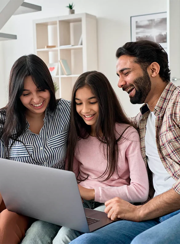 A woman, a man, and a young girl sit together on a couch, smiling and looking at a laptop screen in a modern living room.