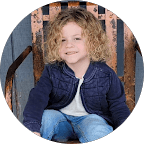 A young child with curly blonde hair, wearing a blue jacket and jeans, sits on a rustic wooden chair and smiles.